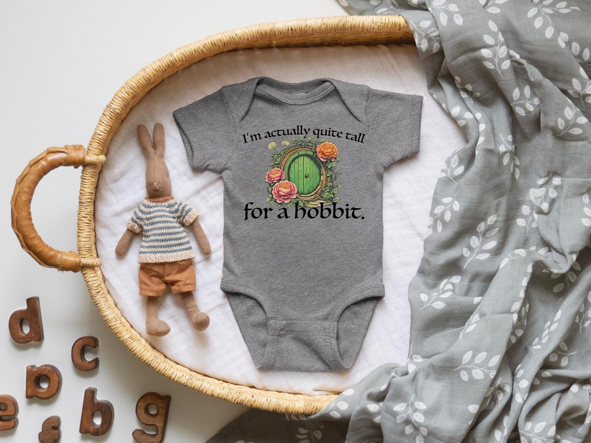 A baby onesie with a humorous text printed on it, placed on a woven basket, alongside a stuffed rabbit and a blanket with a leaf pattern.