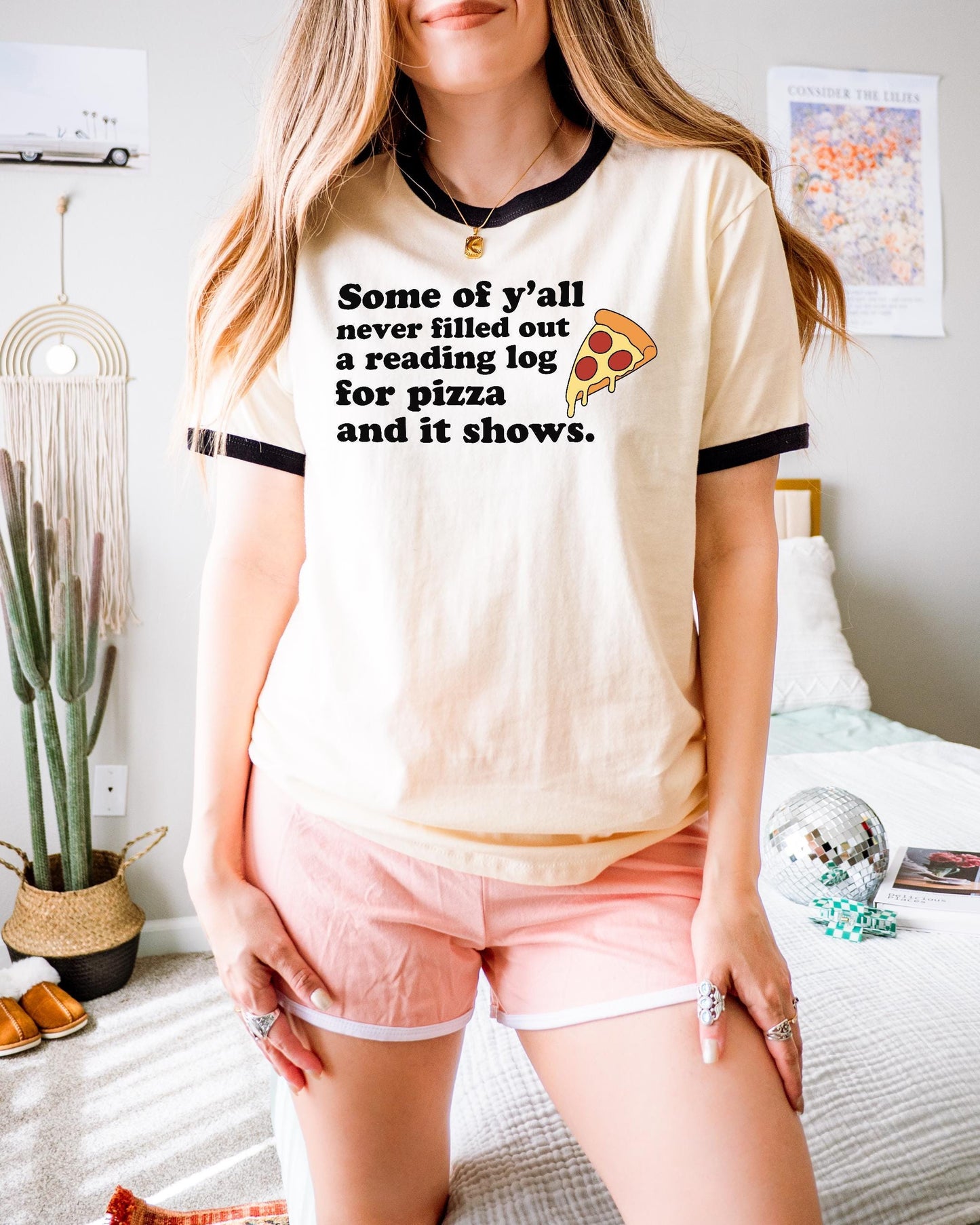 A woman wearing a white t-shirt with a humorous text printed on it, standing in a room with a bed, plants, and a mirror.