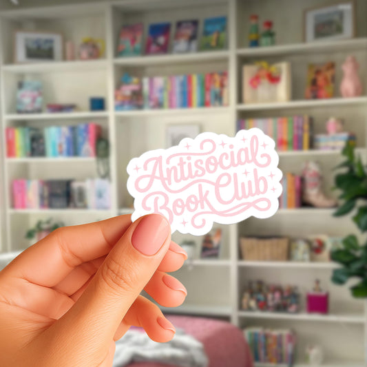 A hand holding a sticker with the text "Antisocial Book Club" in a room with bookshelves filled with books.