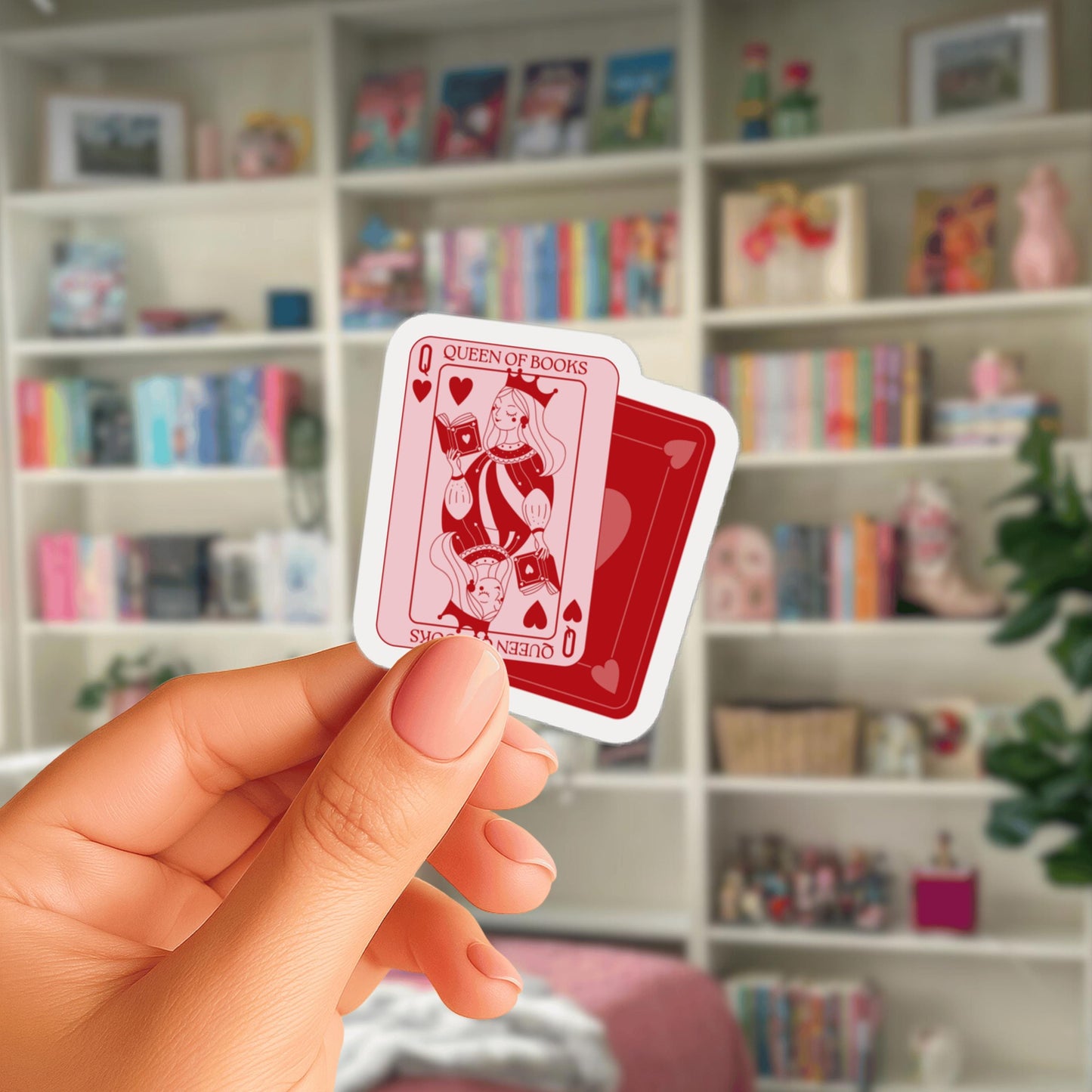 A hand holding a small red and white playing card with a picture of a queen of hearts on it, against a blurred background of bookshelves.