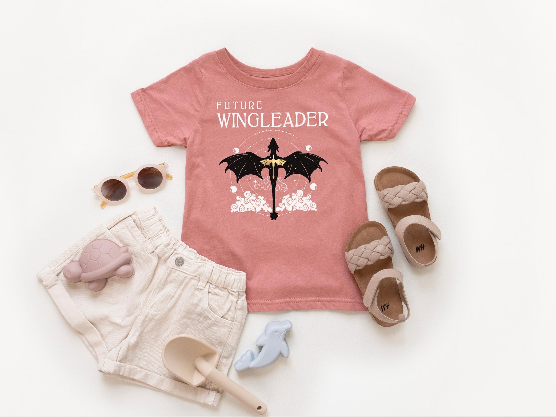 A pink t-shirt with a dragon design, a pair of beige sandals, a pair of sunglasses, and a small toy on a white background.