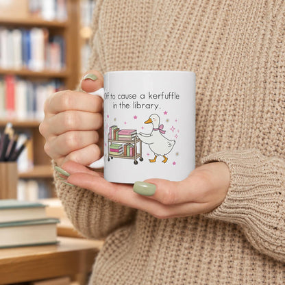 A person is holding a white mug with a cartoon duck and the text "Off to cause a kertffuffle in the library" printed on it.