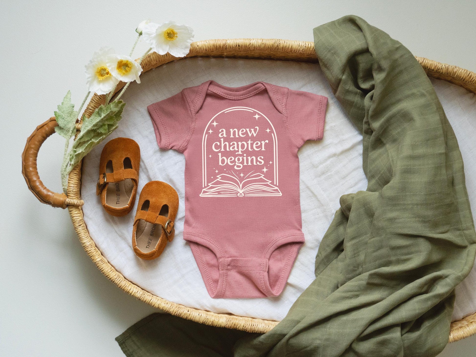 A pink onesie with the text "a new chapter begins" printed on it, along with a pair of brown baby shoes, a green blanket, and white flowers, all arranged on a woven tray.