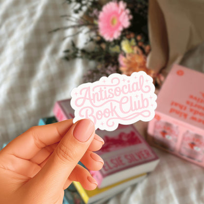 A hand holding a sticker with the text "Antisocial Book Club" in a pink color, with a blurred background featuring books and flowers.