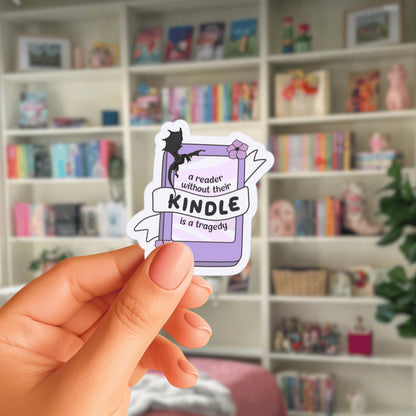 A hand holding a sticker with the text "a reader without their Kindle is a tragedy" against a background of bookshelves filled with various books.