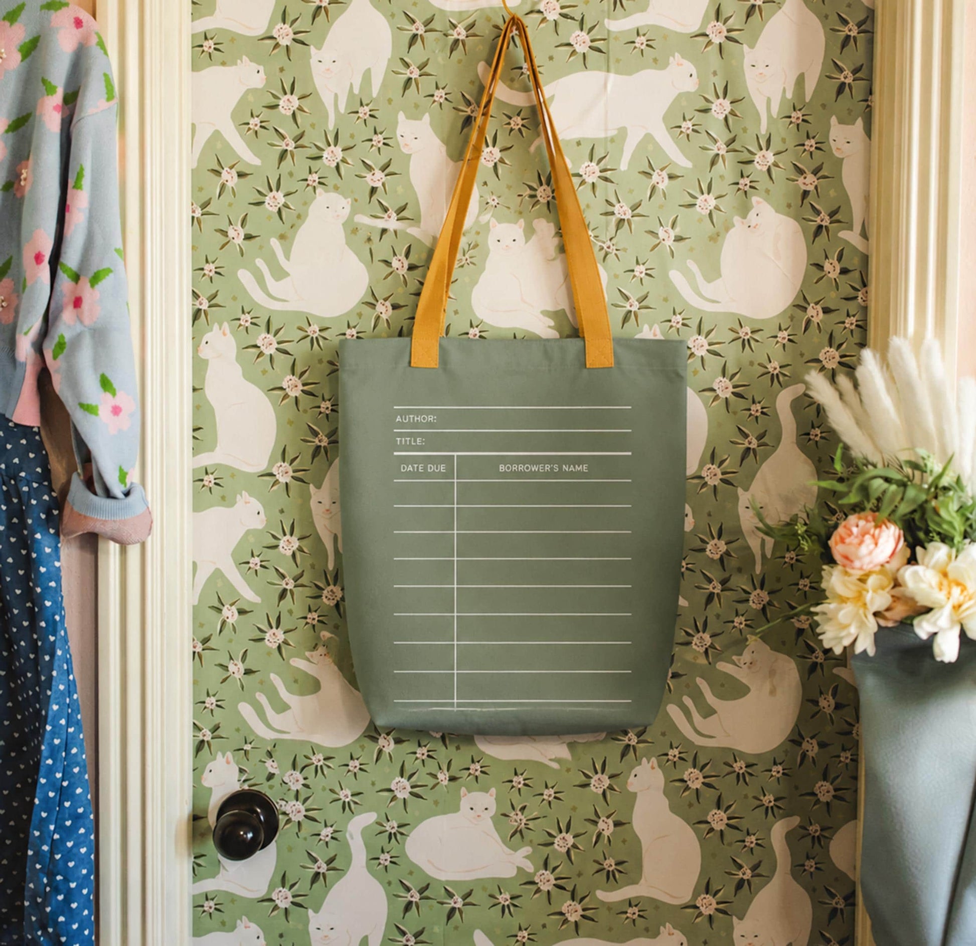 a green tote bag hanging on a wall with a pattern of white cats. The bag has a list of information printed on it, including the title, author, title, and date due. The wall behind the bag has a floral pattern.