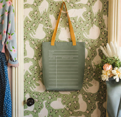 a green tote bag hanging on a wall with a pattern of white cats. The bag has a list of information printed on it, including the title, author, title, and date due. The wall behind the bag has a floral pattern.