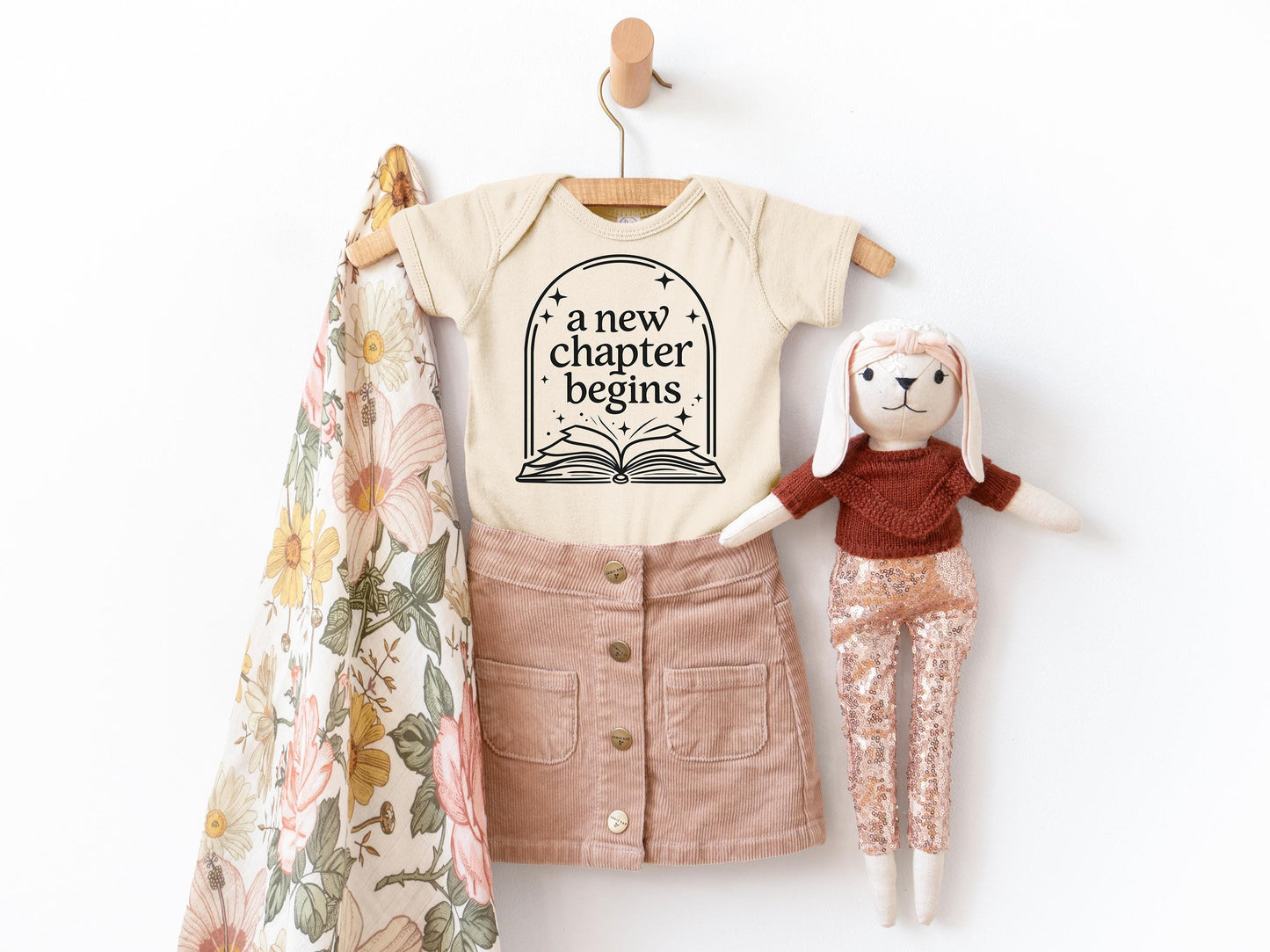 A baby's outfit with a t-shirt and skirt, along with a stuffed rabbit, is displayed on a hanger against a white background.