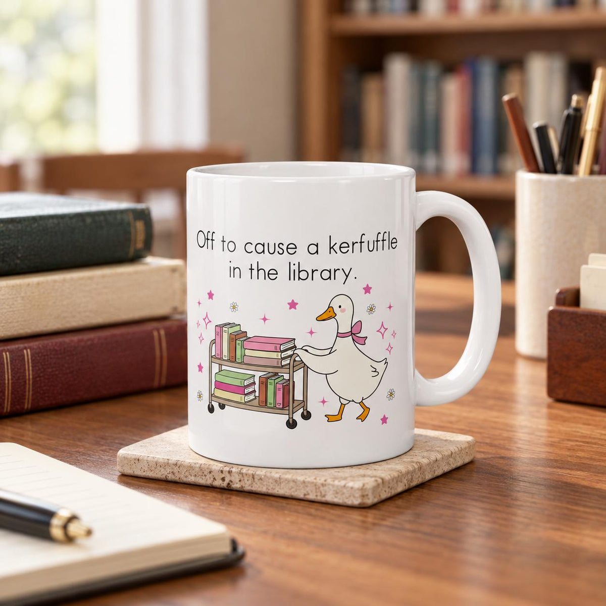 A white mug with a cartoon duck and the text "Off to cause a kerfuffle in the library" printed on it, placed on a wooden surface with a stack of books and a pen nearby.