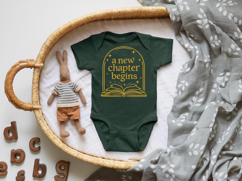 A baby onesie with the text "a new chapter begins" printed on it, placed in a woven basket. Next to the onesie is a stuffed rabbit, and there are wooden letters on the side of the basket.