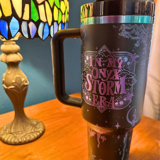 A 40 oz engraved tumbler inspired by Fourth Wing, featuring an Onyx Storm Era design with intricate dragons and fantasy book elements. This Stanley-style cup keeps drinks hot or cold for hours with double-wall insulation. Comes with a handle, sliding lid, and reusable straw—perfect for dragon book lovers and fantasy fans!
