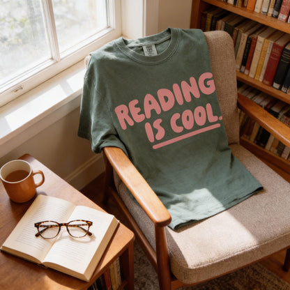 A green t-shirt with the text "Reading is cool" is draped over a wooden chair, with a pair of glasses and a book on a nearby table.