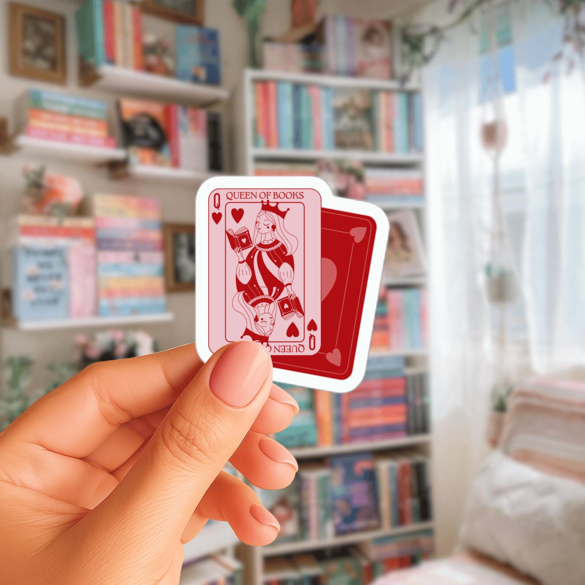 A hand holding a small red and white sticker with a queen of books design, in front of a bookshelf filled with books.