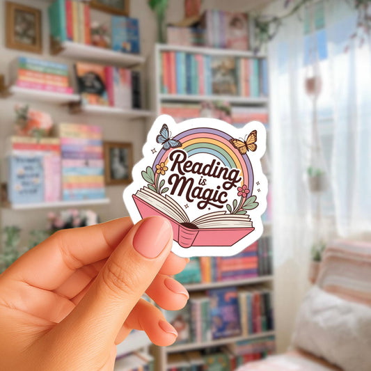 A hand holding a sticker with the text "Reading is Magic" and an illustration of an open book with a rainbow and butterflies.