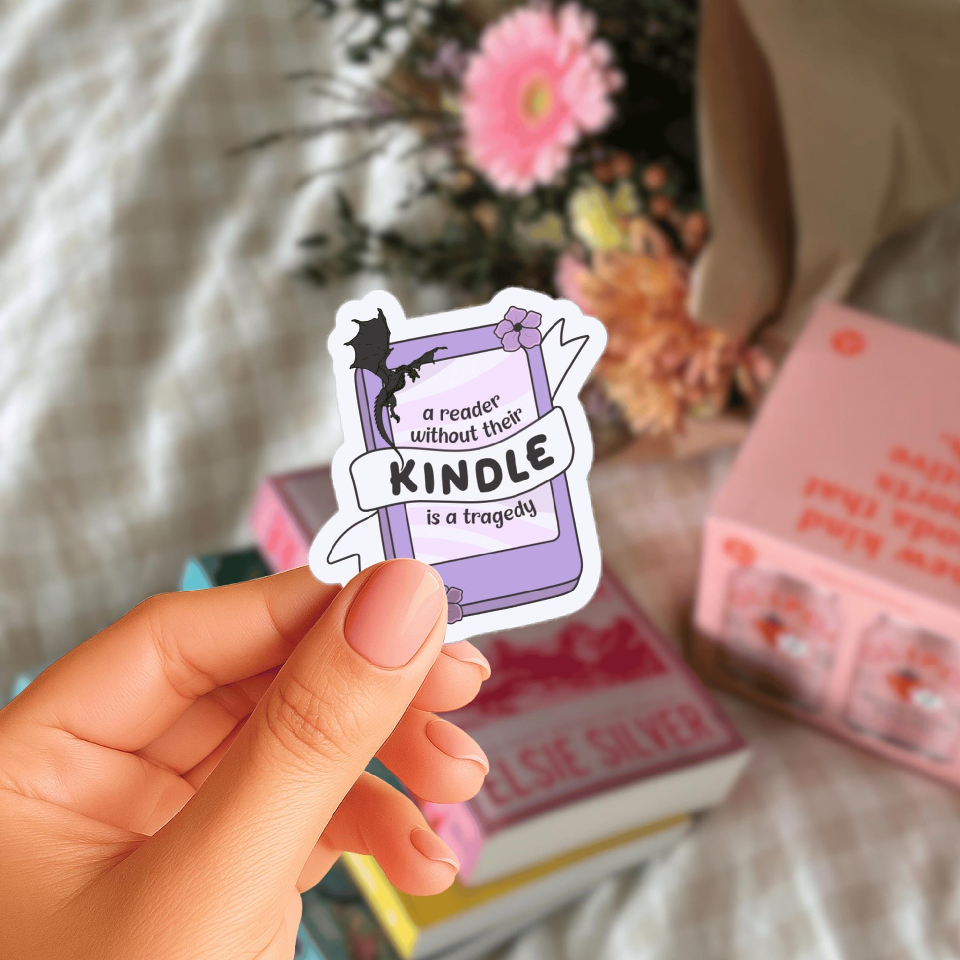 A hand holding a sticker with the text "a reader without their Kindle is a tragedy" against a background of books and flowers.