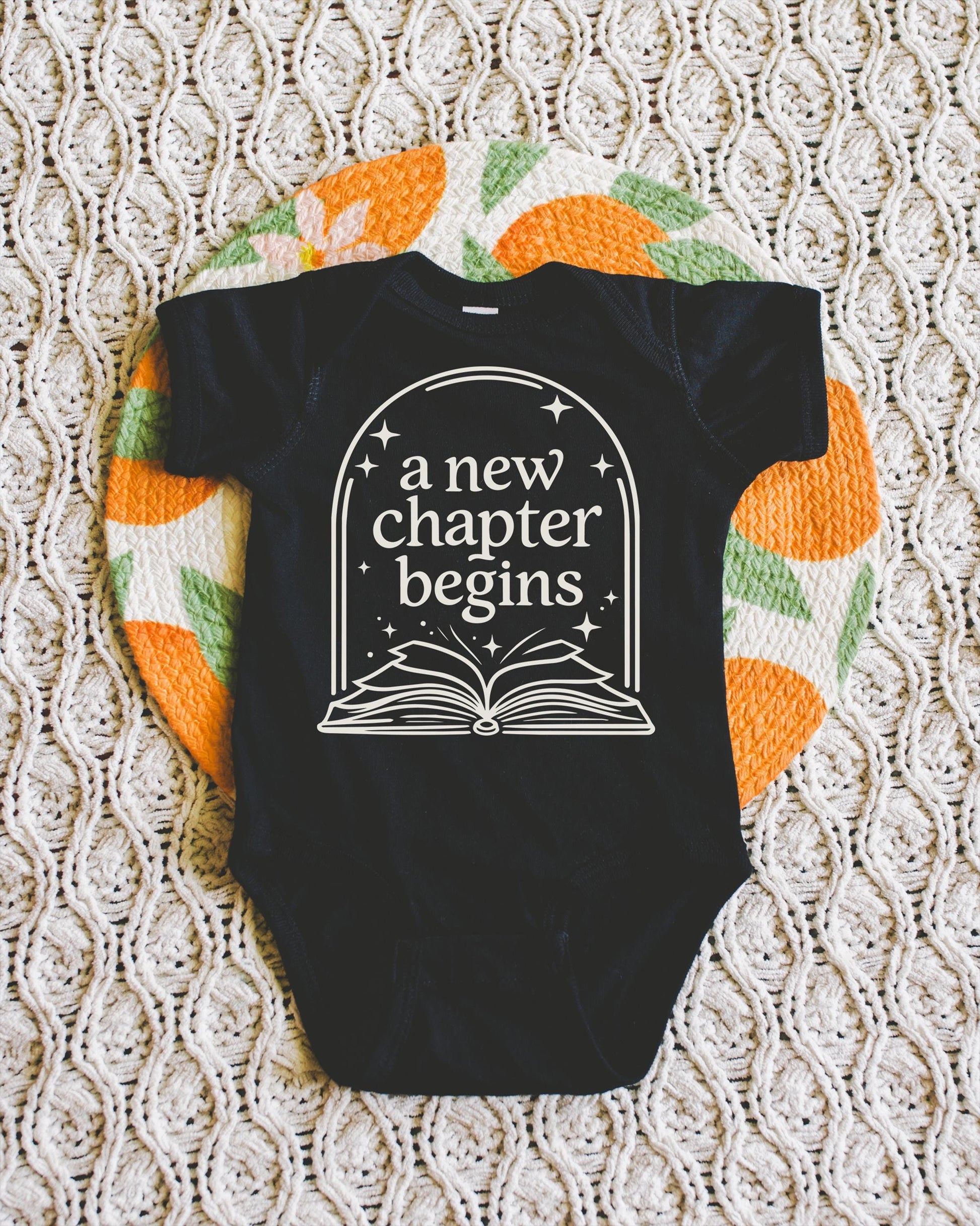 A black baby onesie with the text "a new chapter begins" printed on it, laid out on a white and orange patterned blanket.
