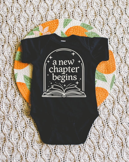 A black baby onesie with the text "a new chapter begins" printed on it, laid out on a white and orange patterned blanket.
