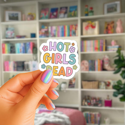 A hand holding a sticker with the text "HOT GIRLS READ" in a colorful, playful font.