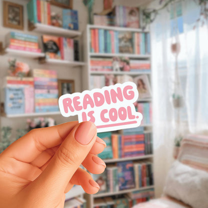 A hand holding a sticker with the text "Reading is cool." in front of a bookshelf filled with books.