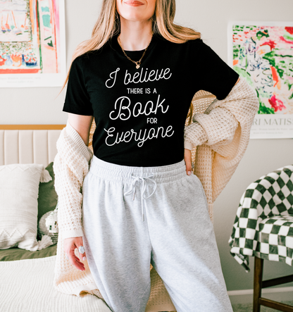 A woman wearing a black t-shirt with white text and a white cardigan stands in a room with colorful artwork on the walls.