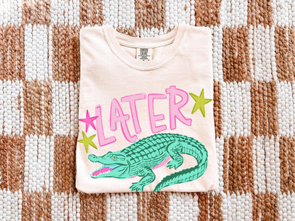 a white t-shirt with a graphic design of a green alligator and pink stars.