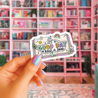 A hand holding a sticker with the text "It's a good day to read a book" against a background of a pink bookshelf filled with books.