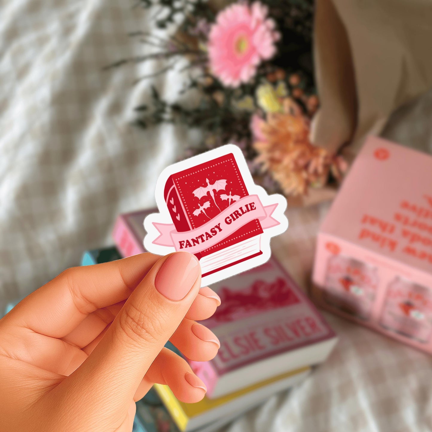 A hand holding a red sticker with the text "FANTASY GIRLIE" on it, with a blurred background of books and flowers.