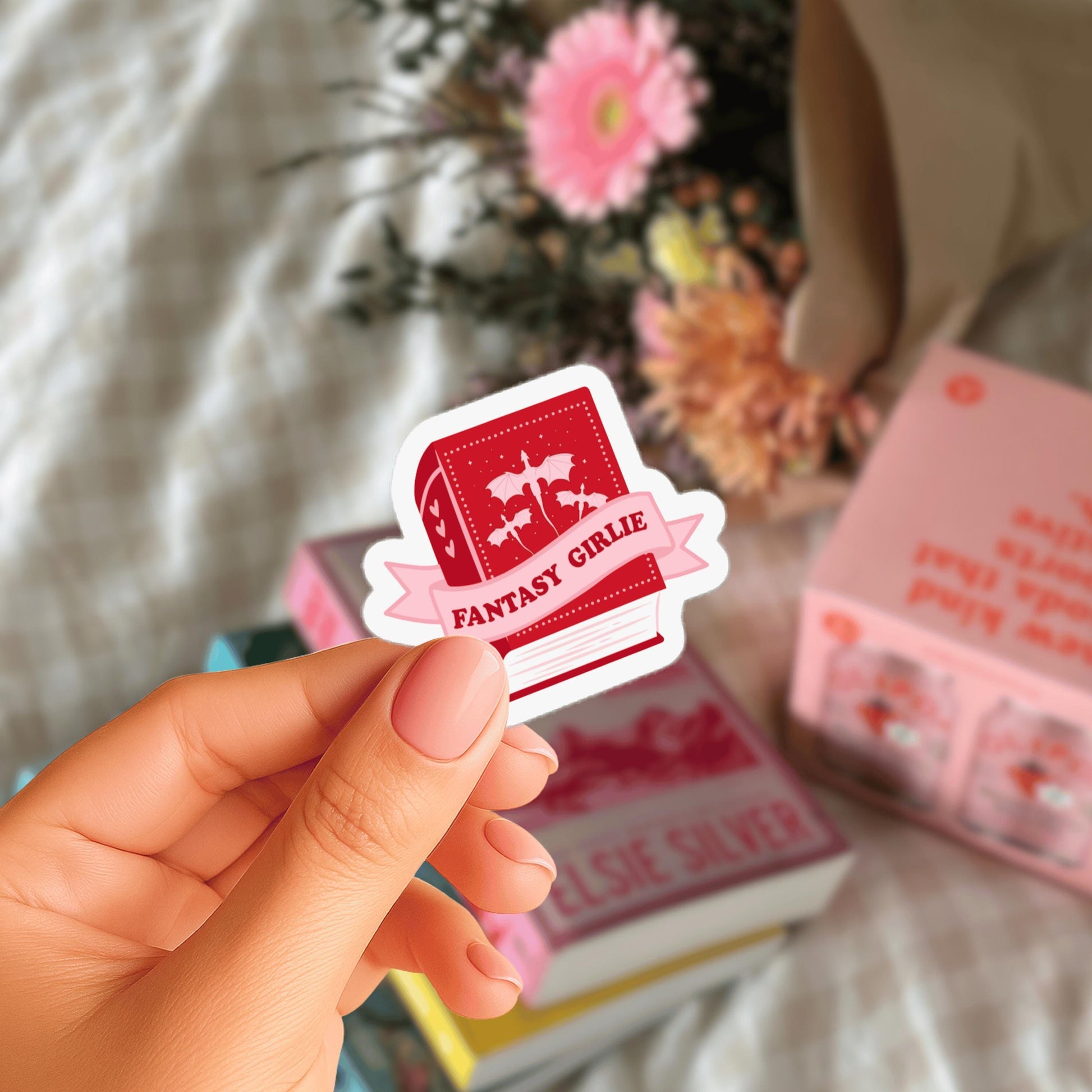 A hand holding a red sticker with the text "FANTASY GIRLIE" on it, with a blurred background of books and flowers.