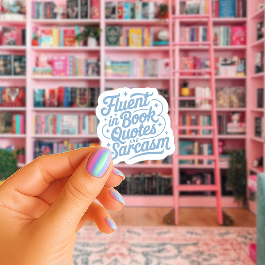 A hand holding a sticker with the text "Flent in Book Quotes and Sarcasm" in a room with pink bookshelves and a pink rug.