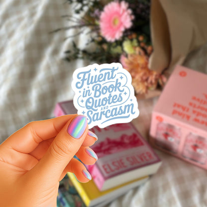 A hand holding a sticker with the text "Flent in Book Quotes and Sarcasm" against a background of books and flowers.