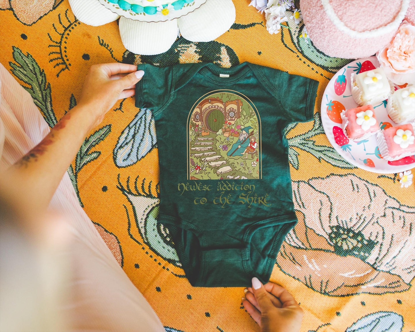 May include: Dark green baby onesie with a whimsical illustration of a hobbit hole and a wizard. The text "Newest Addiction to the Shire" is printed below the graphic. The onesie is displayed on a floral patterned blanket.