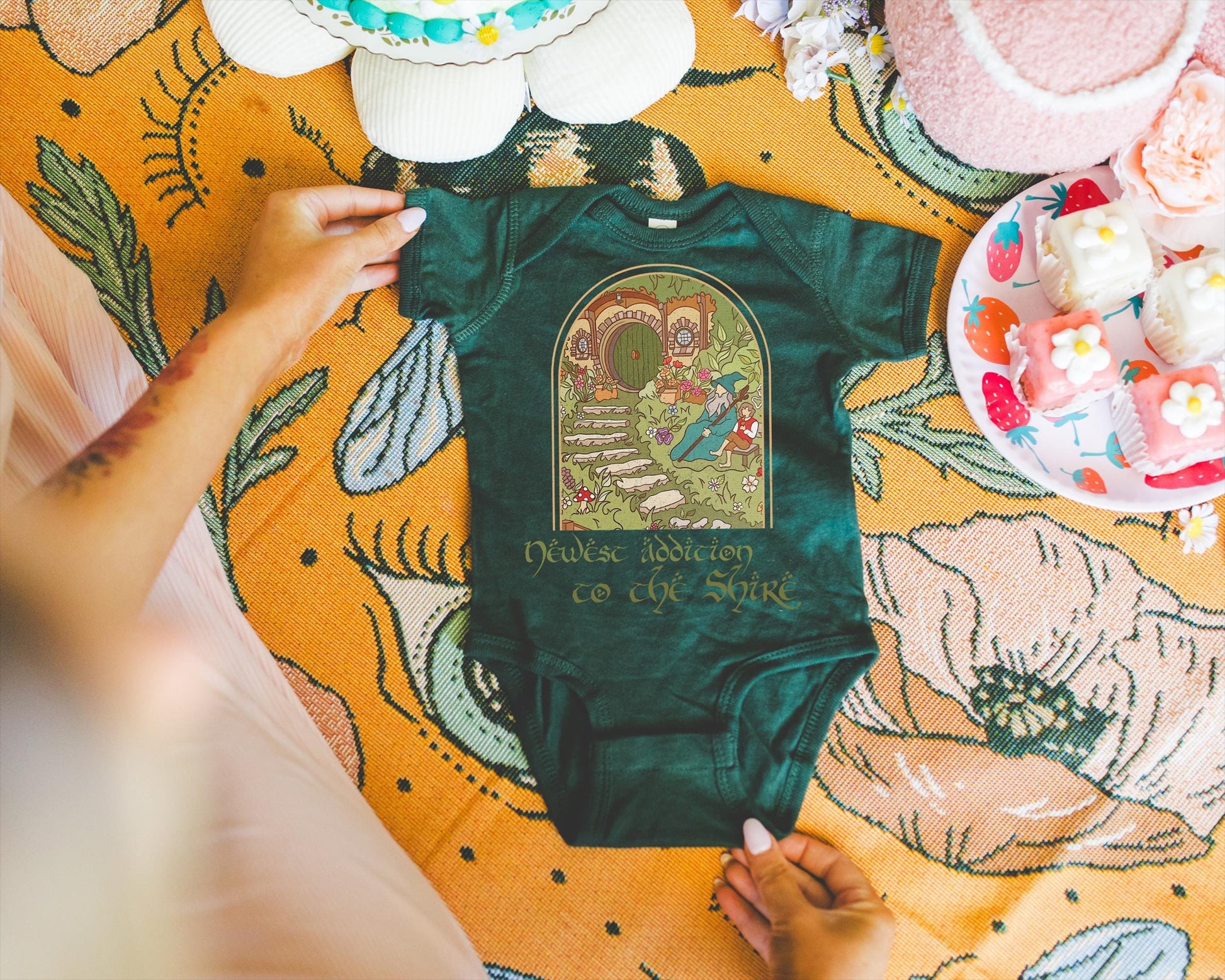 May include: Dark green baby onesie with a whimsical illustration of a hobbit hole and a wizard. The text "Newest Addiction to the Shire" is printed below the graphic. The onesie is displayed on a floral patterned blanket.