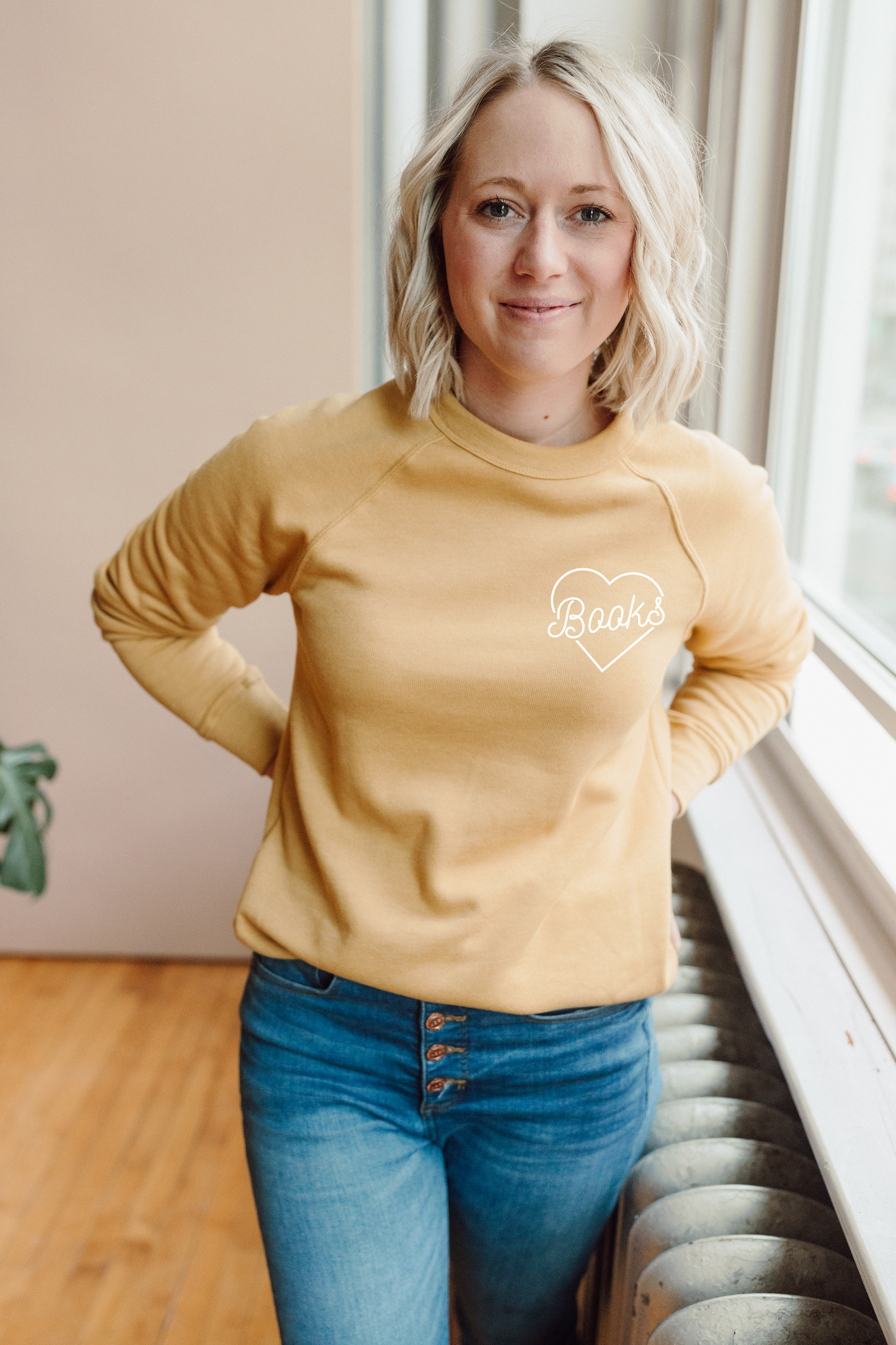 A woman with blonde hair is standing in front of a window, wearing a yellow sweatshirt with the word "Books" printed on it.