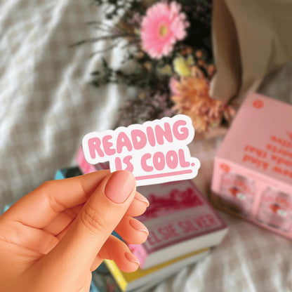 A hand holding a pink sticker with the text "Reading is cool." in front of a blurred background with flowers and books.
