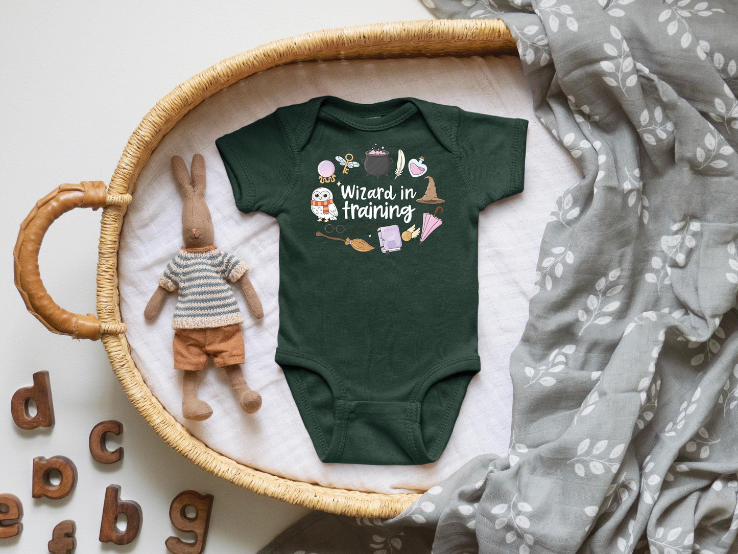 May include: Dark green baby bodysuit with the text "Wizard in training" and whimsical illustrations of a broom, owl, potion, and spell book. The bodysuit is displayed in a woven basket with a stuffed bunny.
