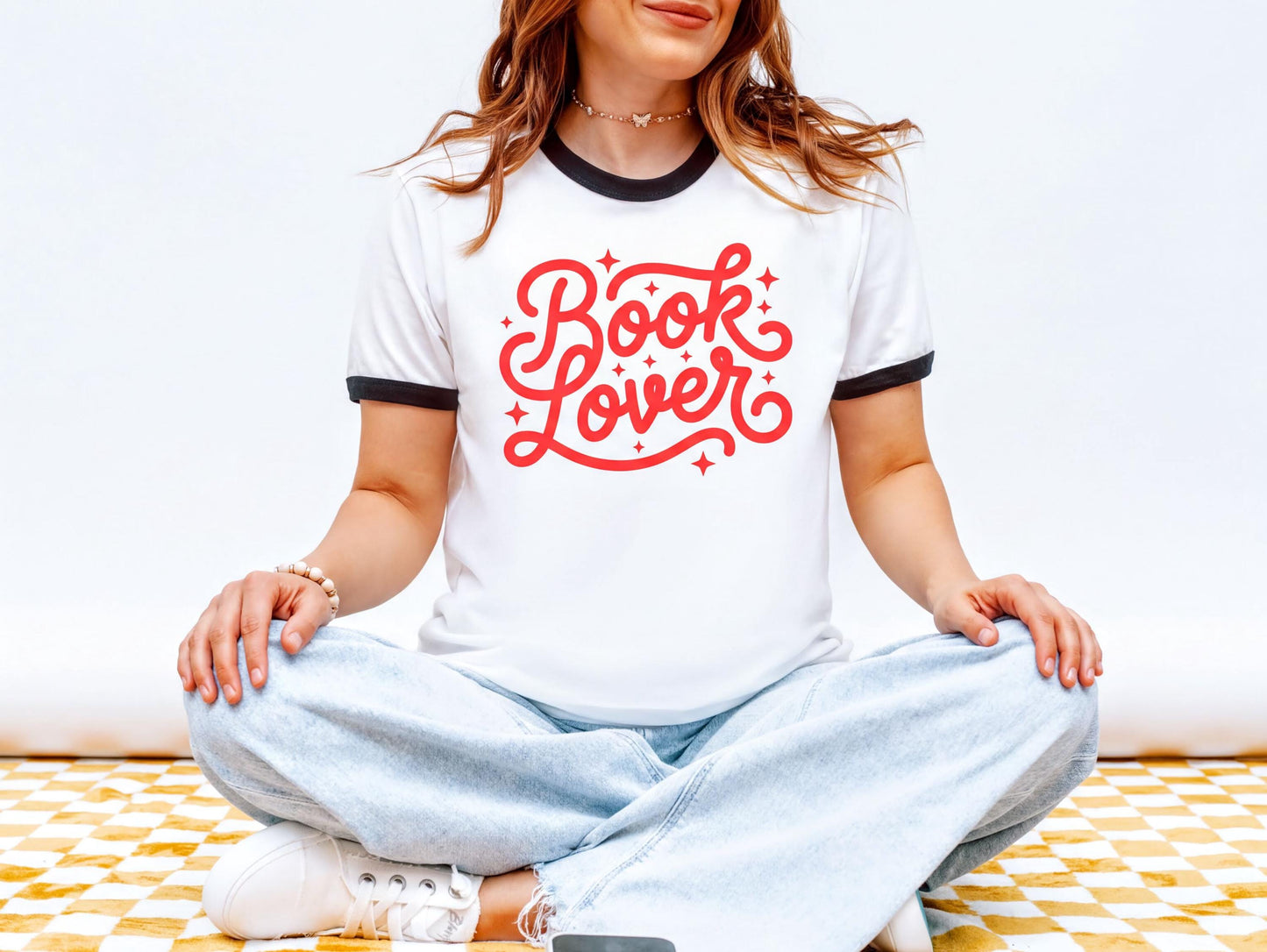 A woman wearing a white t-shirt with red text and jeans is sitting cross-legged on a checkered floor.