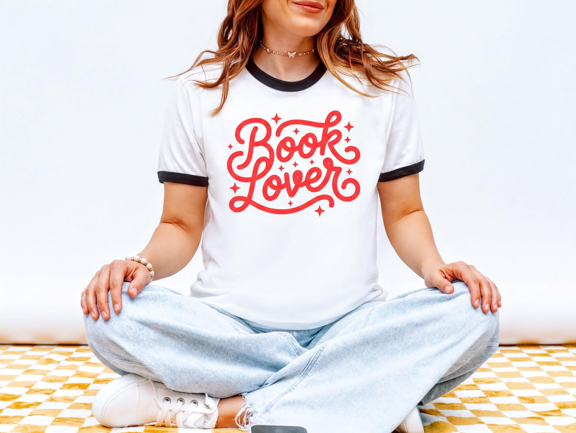 A woman wearing a white t-shirt with red text and jeans is sitting cross-legged on a checkered floor.